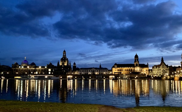 Ein Fluß vor der Silhouette einer Stadt mit Türmen und Kuppeln am Abend, in dem sich Lichter spiegeln