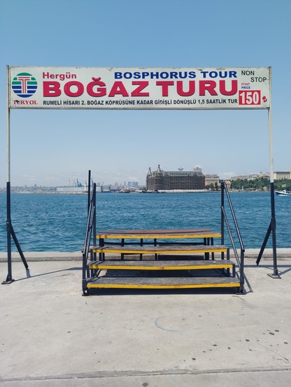 Ein Fähranleger. Im Vordergrund steht eine Treppe mit vier Stufen und Geländer. Darüber steht ein großes Schild, auf dem in türkisch eine Bosporus-Tour angeboten wird. Das Schild ist weiß, die Schrift ist rot. Im Hintergrund ist Wasser, ganz hinten sind Hafenanlagen zu sehen.