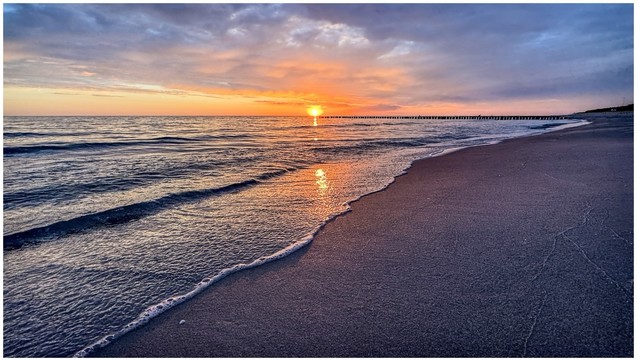 Sunrise over a tranquil beach at the Baltic Sea with gentle waves, reflecting the orange and pink hues of the sky. A pier extends into the water in the distance.