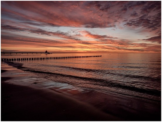 Sunset over a calm beach, with a pier extending into the water and colorful clouds in the sky.