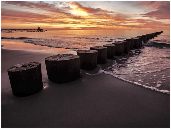 Wooden posts extending into the Baltic Sea with a vibrant sunset in the background and gentle waves washing onto the sandy shore.