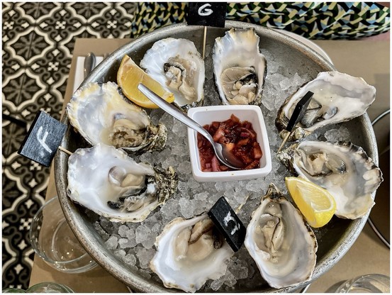 A plate of fresh oysters on a bed of ice garnished with lemon wedges and served with a small container of red sauce, surrounded by small black flag markers.