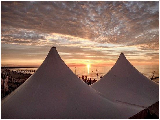 Two white tents by the beach at sunset, with a dramatic sky and the sun setting over the ocean in the background.