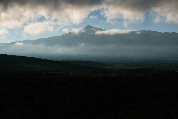 Cloudy morning view at the Sangre de Cristo Mountains. The   mesa is still shrouded in deep, black shadows. The distant mountain peak is clearly outlined against a blue sky and framed by heavy clouds above and whisps of white mist below.