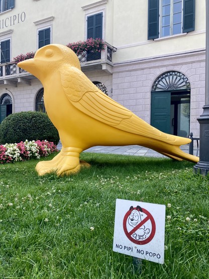 Auf einem Beet vor einer Häuserzeile steht eine überdimensionale gelbe Vogelskulptur
