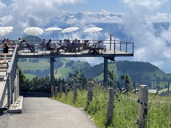Ein Motiv in den Schweizer Alpen. Auf einer von Säulen getragenen Terrasse sitzen Gäste eines Cafés unter weißen Sonnenschirmen.