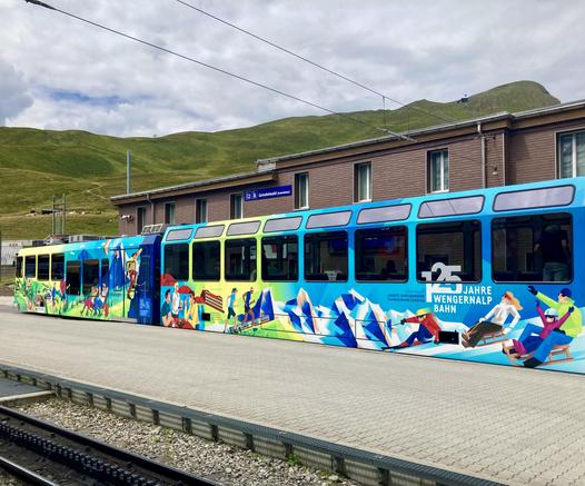 Auf einem Bahnhof hält ein bunt mit touristischen Motiven bemalter Zug, im Hintergrund Berge