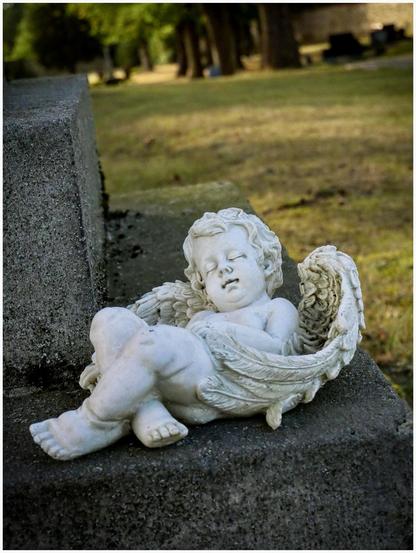 A small marble sculpture of an angelic figure resembling a sleeping baby with wings, resting on a stone surface, surrounded by a grassy area and trees in the background.