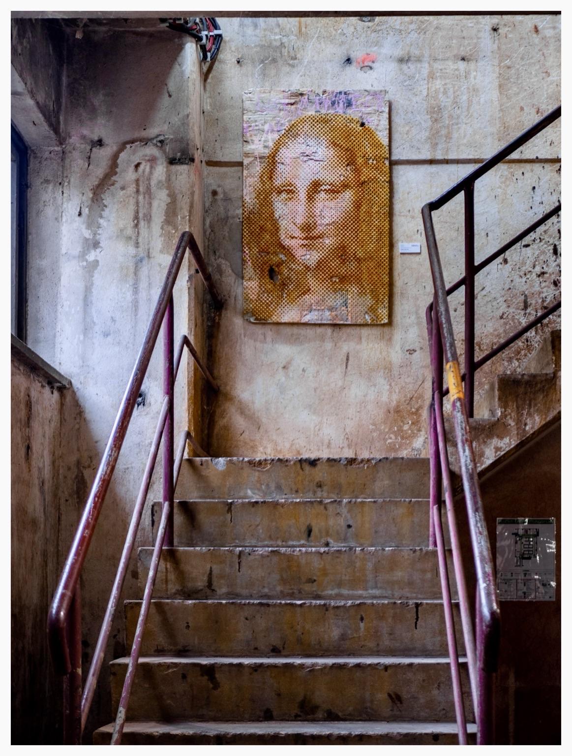 A staircase in an industrial setting with worn steps and rusty railings. On the wall, there is a large, textured artwork resembling the Mona Lisa, featuring a honeycomb pattern and muted colors. The surroundings are characterized by peeling paint and a raw, A staircase in an industrial setting with worn steps and rusty railings. On the wall, there is a large, textured artwork resembling the Mona Lisa, featuring a honeycomb pattern and muted colors. The surroundings are characterized by peeling paint and a raw,