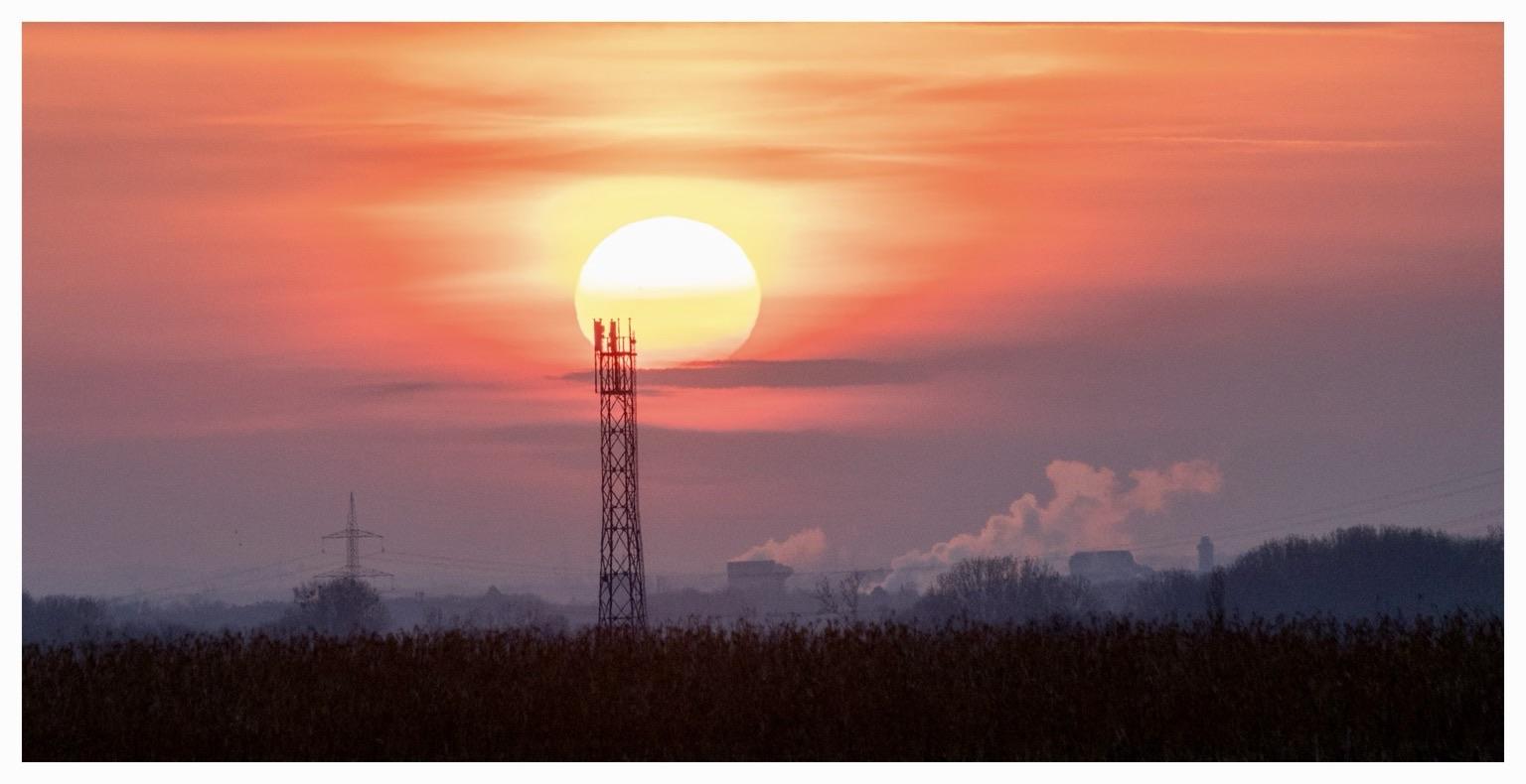 A sunset with a large sun partially obscured by clouds, casting orange and pink hues across the sky. In the foreground, there is a silhouette of a communication tower and smoke rising from industrial buildings in the background. A sunset with a large sun partially obscured by clouds, casting orange and pink hues across the sky. In the foreground, there is a silhouette of a communication tower and smoke rising from industrial buildings in the background.