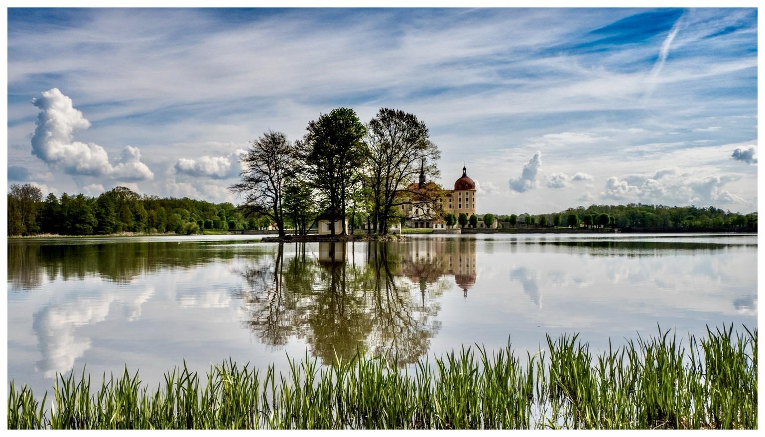 A scenic view of a castle surrounded by lush greenery and trees, reflected in a calm lake. The sky is partly cloudy with a mix of blue and white clouds. Reeds are visible in the foreground. A scenic view of a castle surrounded by lush greenery and trees, reflected in a calm lake. The sky is partly cloudy with a mix of blue and white clouds. Reeds are visible in the foreground.
