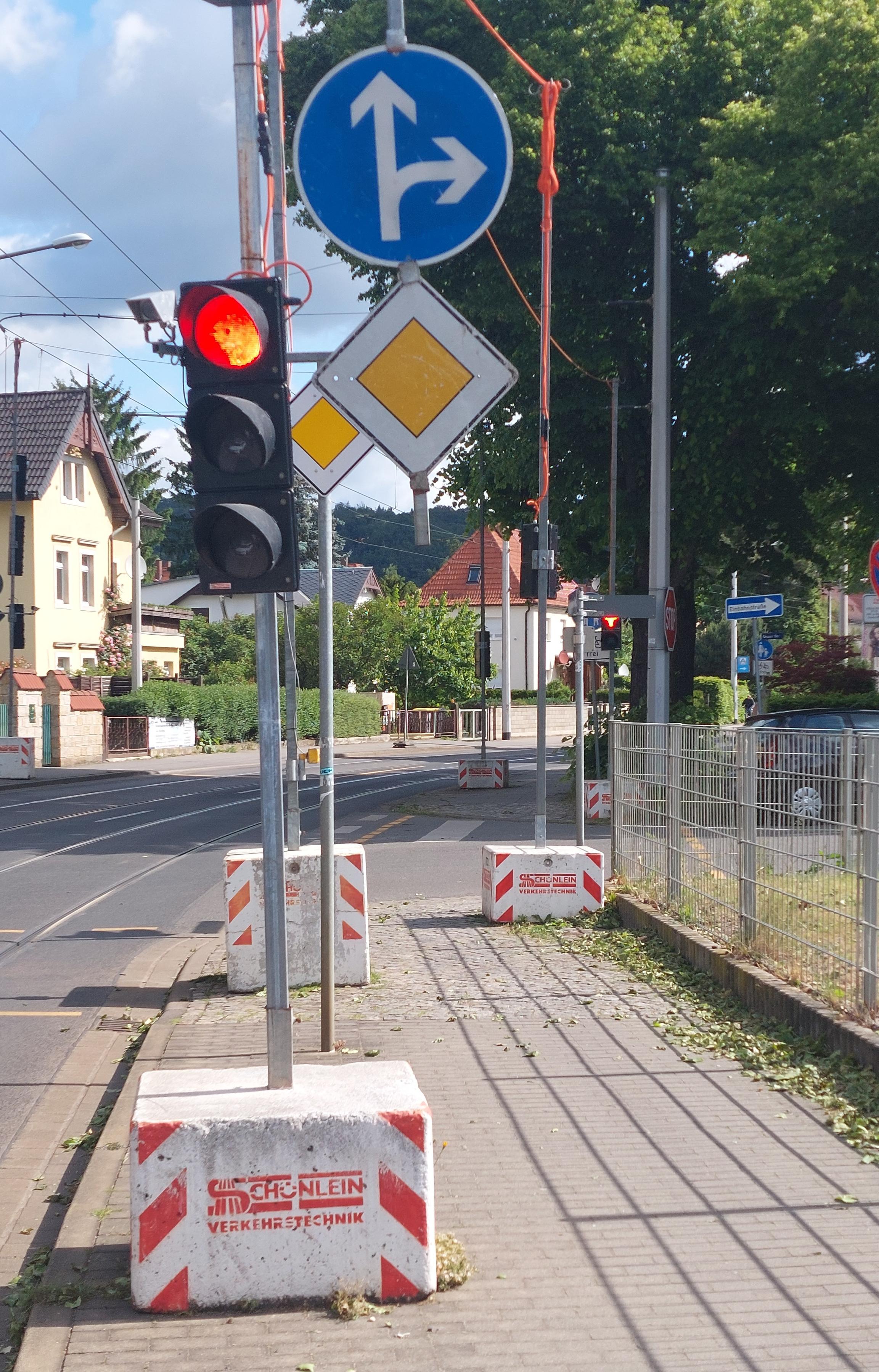 Man sieht einen Fußweg, der auf eine Kreuzung zuläift. Der Fußweg ist vorgestellt mit einer behelfamäßigen Ampel in dicken Betonsockeln und baustellenbedingten Verkehrsschildern. Dazwischen ist nich etwas Platz für Fußgänger. Parallel dazu verläuft die Hauptstraße mit Straßenbahnschienen. Man sieht einen Fußweg, der auf eine Kreuzung zuläift. Der Fußweg ist vorgestellt mit einer behelfamäßigen Ampel in dicken Betonsockeln und baustellenbedingten Verkehrsschildern. Dazwischen ist nich etwas Platz für Fußgänger. Parallel dazu verläuft die Hauptstraße mit Straßenbahnschienen.