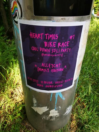 Das Bild zeigt eine Aufkleber-Veranstaltung auf einem Metallbehälter, der auf einer Rasenfläche steht. Der Aufkleber ist in einem dunklen Violettton gehalten und enthält in pinkfarbener Schrift die folgenden Informationen: "HEART TIMES #1 BIKE RACE COOL DOWN SOLI PARTY [@]worldbicyclerelief ALLECAT FAMILY EDITION 31.05.2025 12:30UHR OBJEKT KLEIN A OHNE ANMELDUNG".