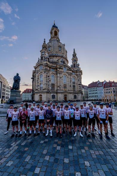 Ein großer Gruppenfoto zeigt eine Gruppe von Radfahrern vor dem historischen Dresdner Frauenkirche. Die Radfahrer tragen weiße Trikots mit roten und schwarzen Mustern und schwarze Radshorts, einige haben auch Helme auf. Sie stehen auf einem gepflasterten Platz, der von der imposanten Barockarchitektur der Frauenkirche dominiert wird.