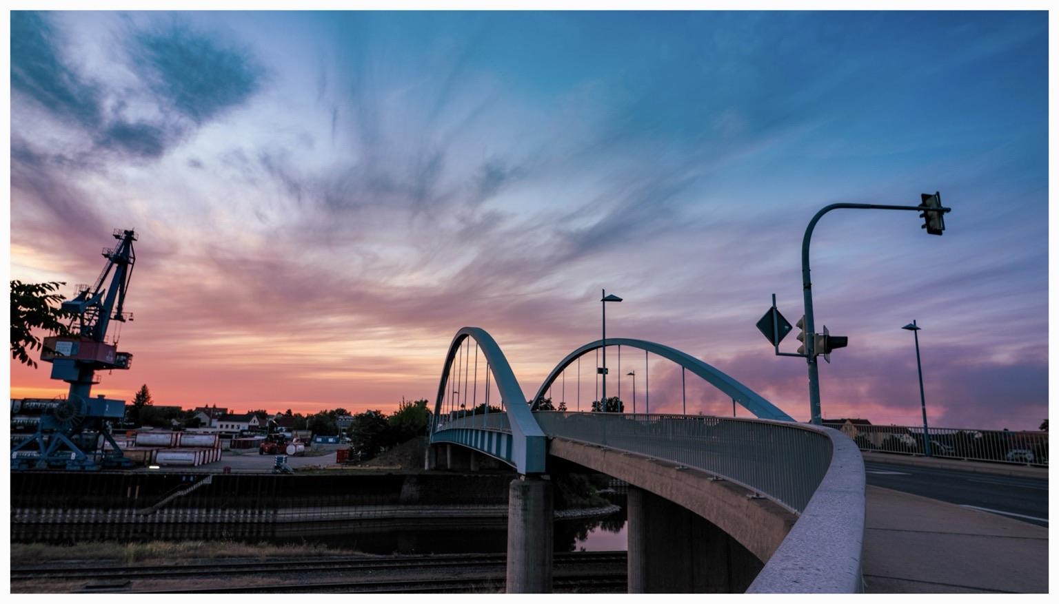 A modern arch bridge spans over a body of water, set against a colorful sunset sky with hues of pink and blue. In the foreground, streetlights and traffic signals are visible, while a blue crane and industrial area can be seen in the background. A modern arch bridge spans over a body of water, set against a colorful sunset sky with hues of pink and blue. In the foreground, streetlights and traffic signals are visible, while a blue crane and industrial area can be seen in the background.