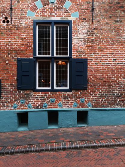 Das Bild zeigt eine Fassade eines historischen Gebäudes mit einer roten Ziegelwand. Im Zentrum der Fassade befindet sich ein großes Fenster mit einem dunkelblauen Rahmen und Gittern, flankiert von zwei dunkelblauen Fensterläden. Über dem Fenster ist ein Bogen mit blauen und roten Ziegeln. Unter dem Fenster befindet sich ein blauer Sitzbalken mit drei archenförmigen Verzierungen. Vor dem Gebäude liegt ein gepflasterter Weg aus roten und braunen Ziegeln. Die Fassade und der Weg vermitteln ein tra…