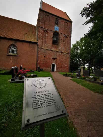 Das Bild zeigt eine alte, rote Backsteinkirche mit einem markanten Turm, der durch eine signifikante Neigung von 5,1939 Grad auszeichnet. Der Turm ist Teil des Protestantischen Kirchengebiets in Suurhusen, Deutschland, und wurde am 17. Januar 2007 für den Guinness World Records certifiziert. Vor der Kirche befindet sich ein gepflasterter Weg, der zu einem Friedhof führt, auf dem mehrere Grabsteine und Blumenarrangements zu sehen sind. Im Vordergrund steht ein silbernes Plaketten, das die Guinne…