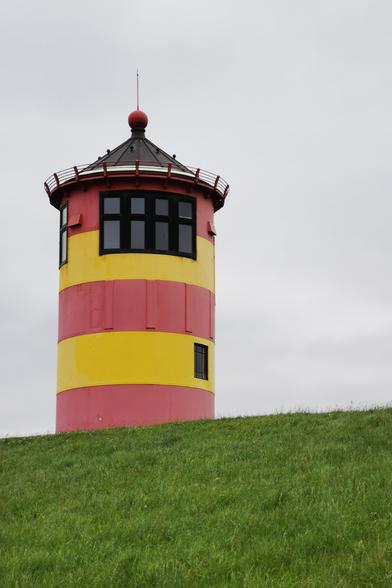 Das Bild zeigt den Pilsumer Leuchtturm, welcher auf einem grünen Deich steht. Das Gebäude ist in leuchtenden Farben gestrichen: rote und gelbe Streifen, die es von der Umgebung abheben. Es hat eine runde Form und ist mit einem Dach mit einem kleinen Turm und einem roten Kugelkopf gekrönt. Die Fenster sind in der oberen Hälfte des Turms angeordnet und haben schwarze Rahmen. Der Himmel ist bedeckt und grau, was dem Bild eine ruhige Atmosphäre verleiht.