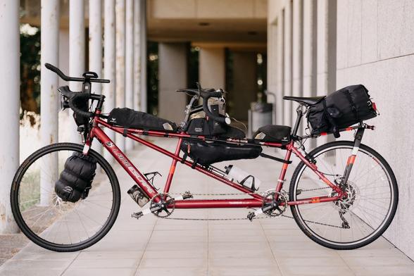 Das Bild zeigt ein rotes Doppelrad, das an einer Wand mit Säulen geparkt ist. Das Rad ist mit verschiedenen Gepäckstücken ausgestattet, darunter eine große schwarze Tasche am Lenker, eine Tasche am Sattel und eine große schwarze Tasche am Hinterrad. Es gibt auch eine kleine Tasche unter dem Sattel und eine Wasserflasche am Rahmen. Das Rad hat eine robuste Konstruktion mit einem Kettenantrieb und Disc-Remissen an beiden Rädern. Die Umgebung ist ein gepflasterter Bereich mit Säulen, die auf eine …