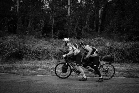 Das Bild zeigt zwei Radfahrer auf einem Fahrrad, das eine tandemkonfiguration hat. Beide tragen Helme und sportliche Kleidung, die für Radfahren geeignet ist. Der Radfahrer im Vordergrund trägt eine kurze Hose und ein ärmelloses Top, während der Radfahrer im Hintergrund eine langärmelige Jacke und eine Hose trägt. Beide haben Rucksäcke und zusätzliche Gepäckstücke an ihrem Fahrrad befestigt, was darauf hindeutet, dass sie möglicherweise auf einer längeren Radtour unterwegs sind.

Sie fahren auf…