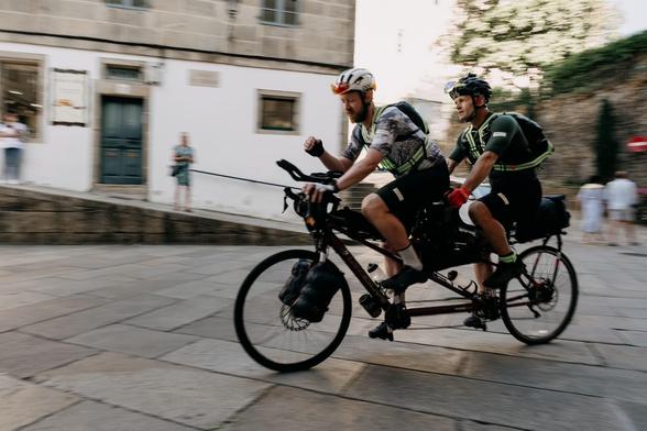 Zwei Männer fahren auf einem Tandemrad durch eine gepflasterte Straße in einer historischen Stadt. Der Fahrer im Vordergrund trägt einen weißen Radhelm und ein gestreiftes Radshirt, während der Ruderer im Hintergrund einen schwarzen Helm und ein grünes Radshirt trägt. Beide sind mit Rucksäcken und Handschuhen ausgestattet. Der Radweg ist von einem Gebäude mit einer weißen Fassade und einem blauen Tor begrenzt, und im Hintergrund sind weitere Personen zu sehen, die entweder beobachten oder weite…