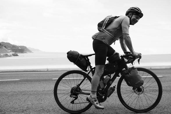 A black and white picture of a cyclist with a loaded bike. The cyclist is travelling in a standing position.