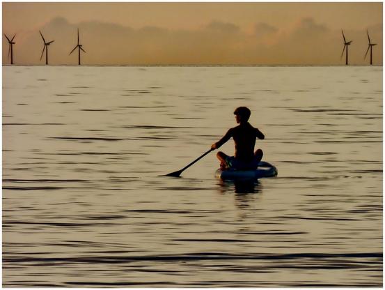 A child paddles on a small inflatable raft in a calm body of water during sunset, with wind turbines visible in the distance against a soft, colorful sky.