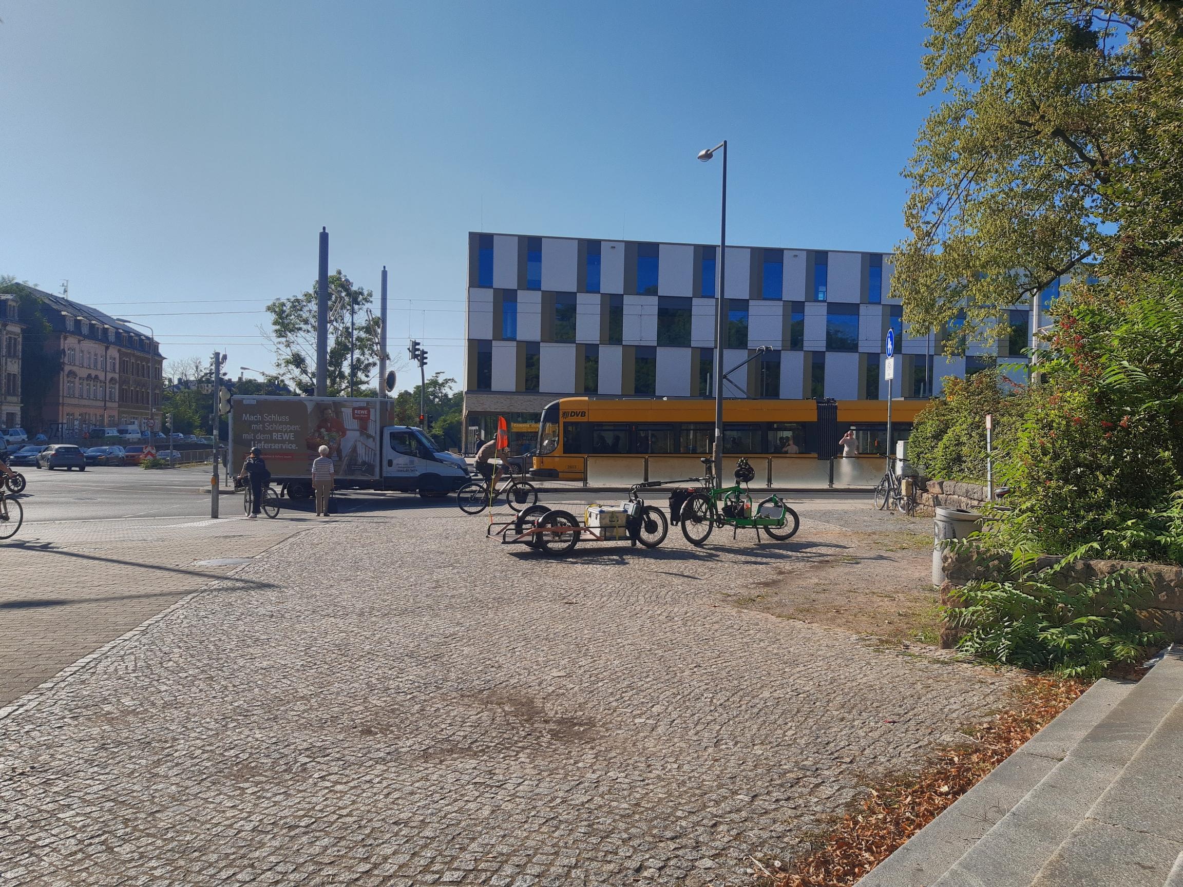 Ein Bullitt Lastenrad steht mit einem Carla Cargo Anhänger auf einem gepflasterten Platz. Dahinter sieht man eine Straße mit Kfz und eine Straßenbahn der Dresdner Verkehrsbetriebe und vereinzelt Personen. Das Wetter ist sonnig.