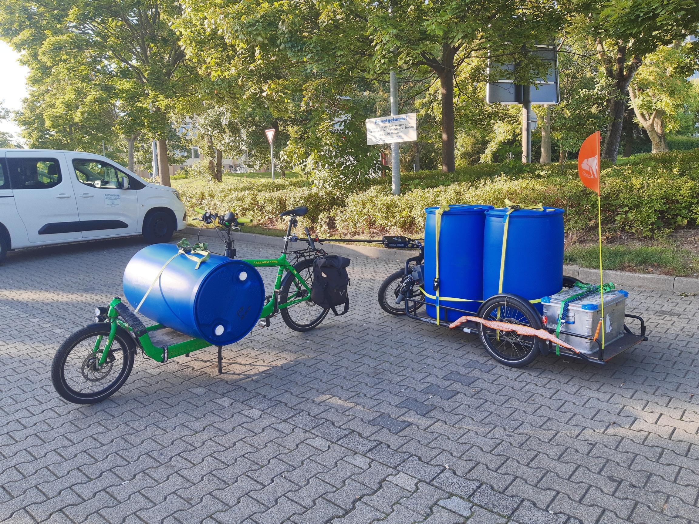 Ein grünes Bullitt Lastenrad mit einem blauen 220l Regenfass quer auf der Ladefläche. Dahinter der zugehörige Carla Cargo Anhänger mit zwei weiteren stehenden 220l Tonnen und einer Alukiste.