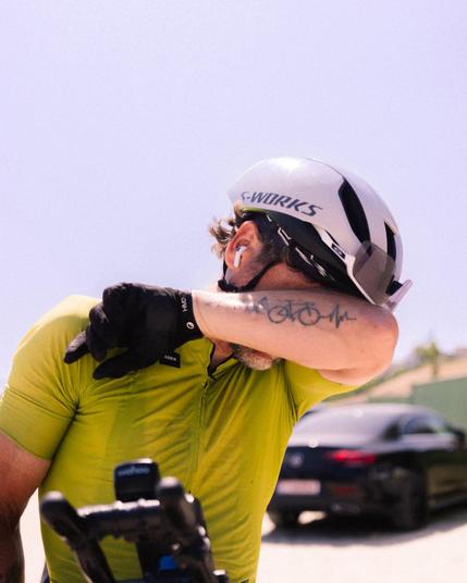 A man in a bright green cycling T-shirt and a white safety helmet with the words "S-WORKS" on it is wiping sweat from his forehead. He's wearing black cycling gloves and white earbuds. On his arm is a tattoo of a bicycle and a heart-shaped pattern. A black sports car and a blue sky can be seen in the background.