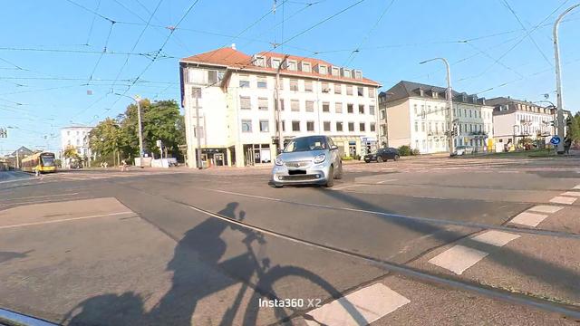 Das Bild zeigt eine Stadtstraße mit einem silbernen Auto, das gerade eine Ampel überquert. Im Vordergrund ist ein Radfahrer zu sehen, dessen Schatten auf der Straße fällt. Das Auto fährt direkt auf den Radfahrenden zu. Im Hintergrund befinden sich mehrstöckige Gebäude mit typisch europäischen Architekturmerkmalen, wie z.B. Fassaden mit Fenstern und roten Dächern. Die Straße ist mit Straßenbahnschienen versehen, und über der Straße verlaufen elektrische Leitungen für die Straßenbahn. Der Himmel …