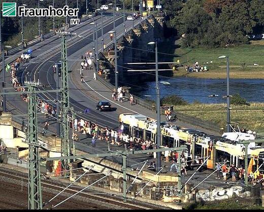 Zu sehen ist das Bild einer Verkehrsbeobachtungskamera, welche auf die Dresdner Marienbrücke blickt. Darauf sperrt eine Menschenkette symbolisch die Straßenbahngleise ab damit die Straßenbahnen freie Fahrt haben und nicht mit den Autos im Stau stehen müssen. 