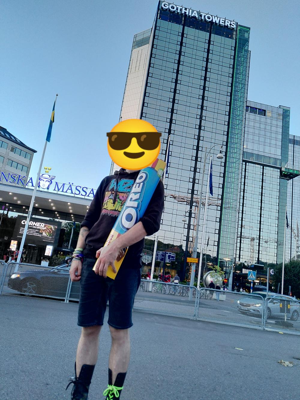 Eine Person, die mit einem Sonnenbrillen Emoji zensiert wurde, steht in einer Stadt auf einem Platz. In der Hand hält die Person eine ca 1 Meter lange Marabou Oreo Schokopackung.
