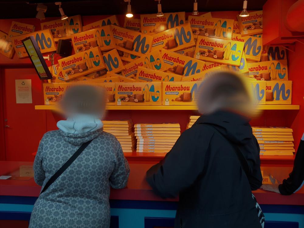 Zwei zensierte Personen stehen vor einer Losbude, in der es riesige Marabou Schokoladentafeln zu gewinnen gibt. Die Tafeln sind an der Wand der Losbude gestapelt.