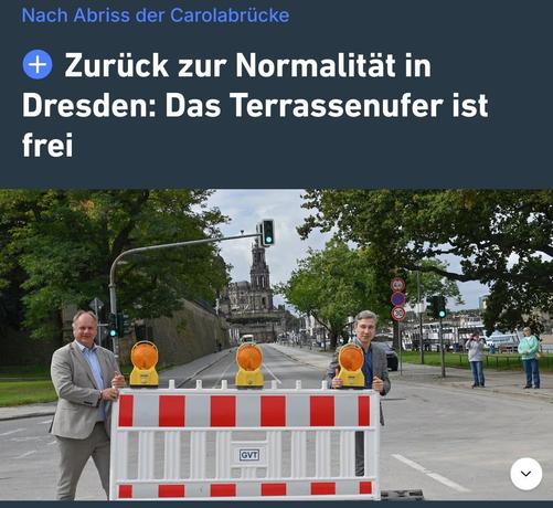 Das Bild zeigt eine Straße in Dresden, Deutschland, nach dem Abriss der Carolabrücke. Im Vordergrund stehen zwei Männer vor einem roten und weißen Bauzaun mit gelben Warnlichtern. Sie scheinen zufrieden und geben ein Zeichen des Erfolgs. Im Hintergrund ist die Elbe zu sehen, sowie die historische Kathedrale mit ihrem markanten Turm, die ein bekanntes Wahrzeichen Dresdens ist. Die Straße ist frei und die Umgebung wirkt normal, was durch den Text "Zurück zur Normalität in Dresden: Das Terrassenuf…