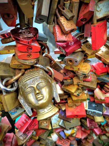 Das Bild zeigt eine Nahaufnahme einer großen Ansammlung von Liebesschlössern, die an einem Geländer befestigt sind. Die Schlösser sind in verschiedenen Größen und Farben, wobei Rot und Gold vorherrschend sind. Im Vordergrund befindet sich ein goldenes Schloss in Form eines Buddha-Gesichts. Mehrere andere Schlösser, einige mit Gravuren, sind in der gesamten Komposition verteilt.