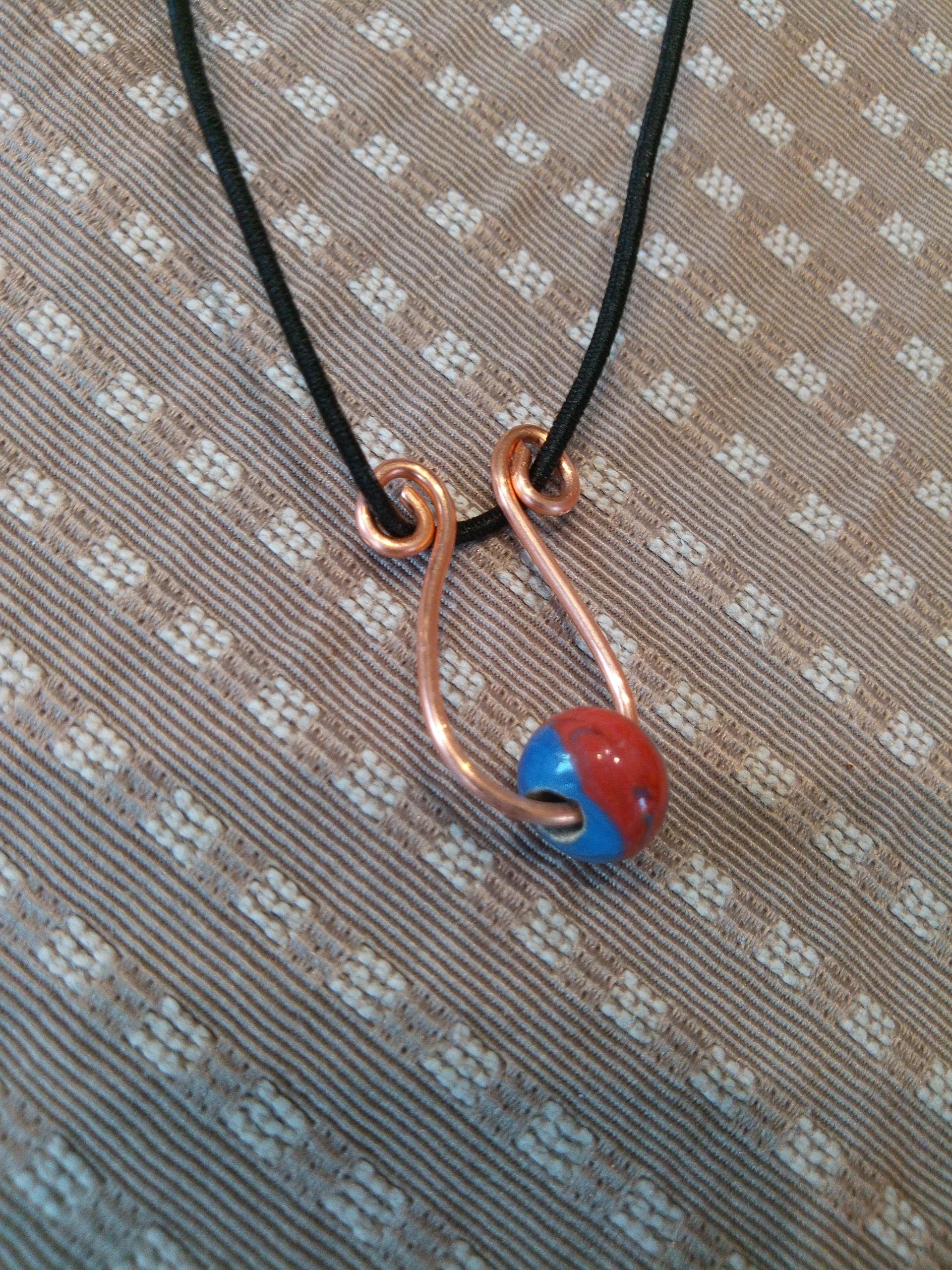 Auf einem Deckchen liegt ein aus Kupferdraht gebogener Anhänger mit einer blauroten Glasperle, aufgefädelt auf schwarzes Lederband.
