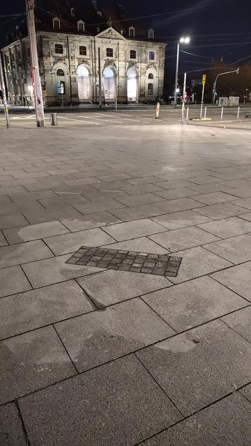 Das Bild zeigt einen leeren Platz bei Nacht. Der Platz ist mit großen quadratischen Betonplatten gepflastert, die in einem regelmäßigen Muster angeordnet sind. Im Hintergrund befindet sich das Blockhaus mit einer Fassade aus Sandstein, das symmetrisch aufgebaut ist und mehrere Fenster und Türen aufweist. Ein Straßenlicht wirft einen Schatten auf die Platten. Im Vordergrund befindet sich schräg zu den Platten eine kleine Pflasterfläche. 