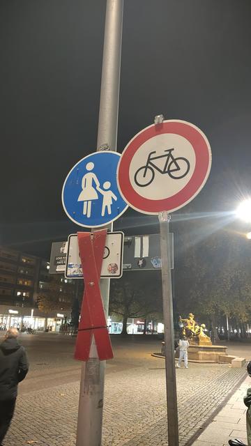 Das Bild zeigt eine Straßenszene bei Nacht. In den Vordergrund ragt ein metallener Mast hervor, an dem mehrere Verkehrsschilder befestigt sind. Oben ist ein Schild mit einem weißen Fahrrad auf rotem Hintergrund zu sehen, das einen Fahrverbot für Fahrräder anzeigt. Daneben befindet sich ein blaues Schild mit einer Abbildung einer erwachsenen und einer kindlichen Figur, welches auf einen Gehweg hinweist. Darunter ist die verdeckte Radfreigabe für den Gehweg. Im Hintergrund sind Gebäude, Straßenbe…