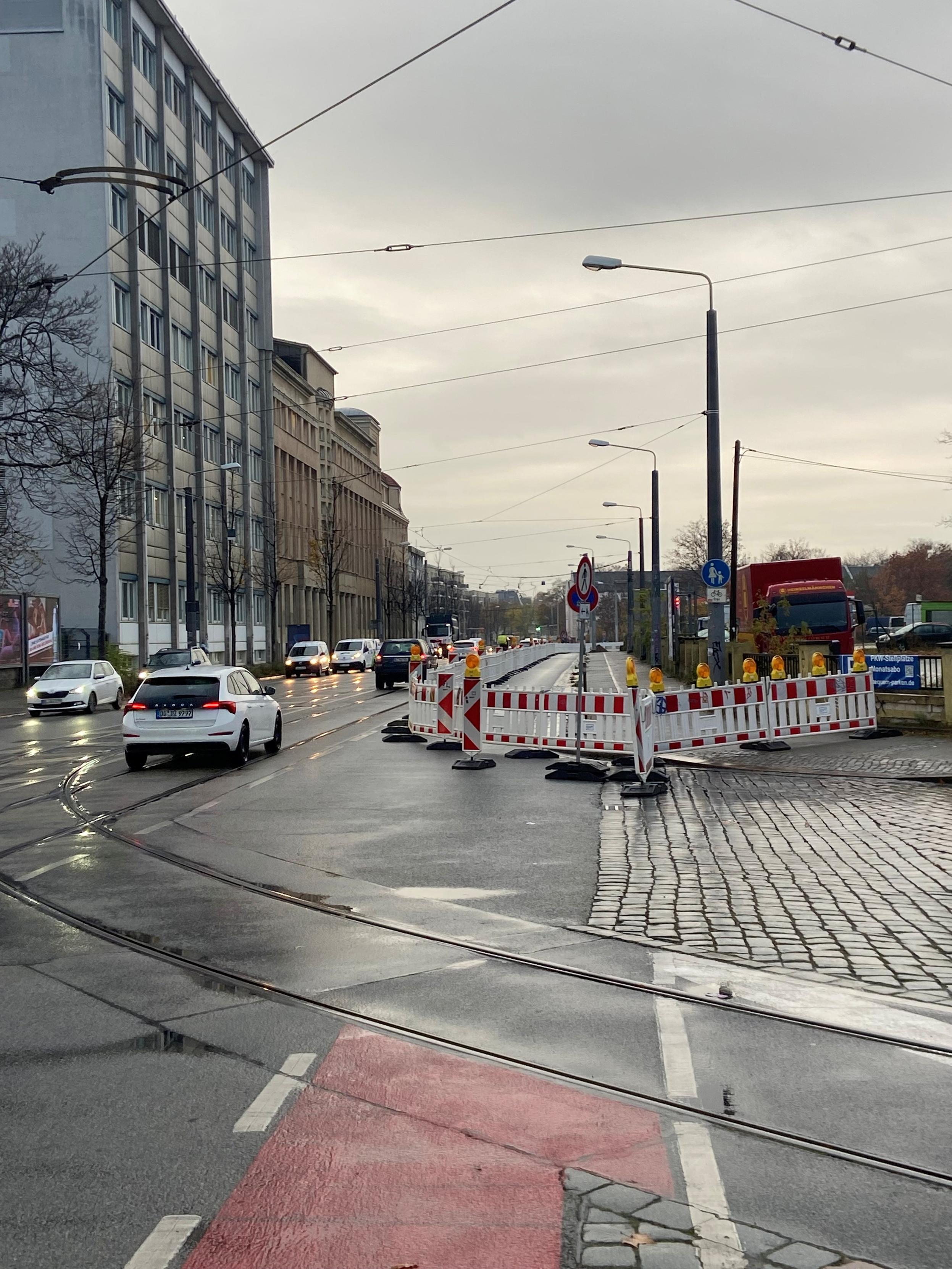 Breite stark befahrene Straße mit Straßenbahn-Schienen in der Mitte. Die rechte Fahrbahn und der Fußweg sind über eine größere Strecke mit Bauzäunen versperrt. Ein Verkehrsschild verbietet das Weitergehen von Fußgängern.