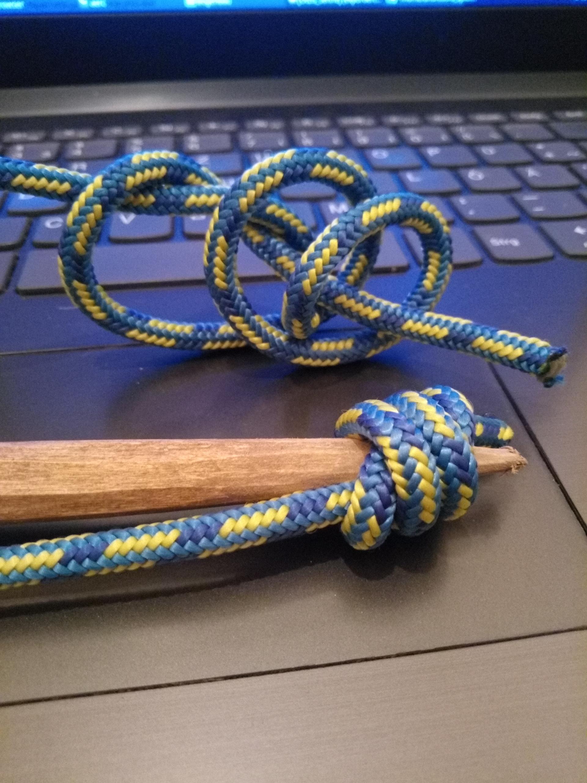 Ein zugespitztes Holz trägt einen Knoten, der sich zusammenzieht. Dahinter der lose gewundene Knoten in blau Reep, damit man die Konstruktion erkennen kann. Alles liegt auf einer grauen Laptoptastatur.