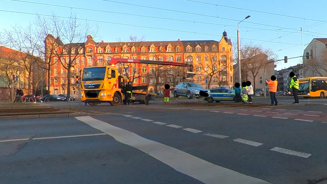 Ein orangenerAbschleppwagen hebt einen silbernen PKW aus dem Gleisbett. Ringsum stehen verschiedene Personen ein Polizeiwagen und ein gelber Bus. Im Hintergrund sieht man eine von der Abendsonne beschienene Hausfassade. 