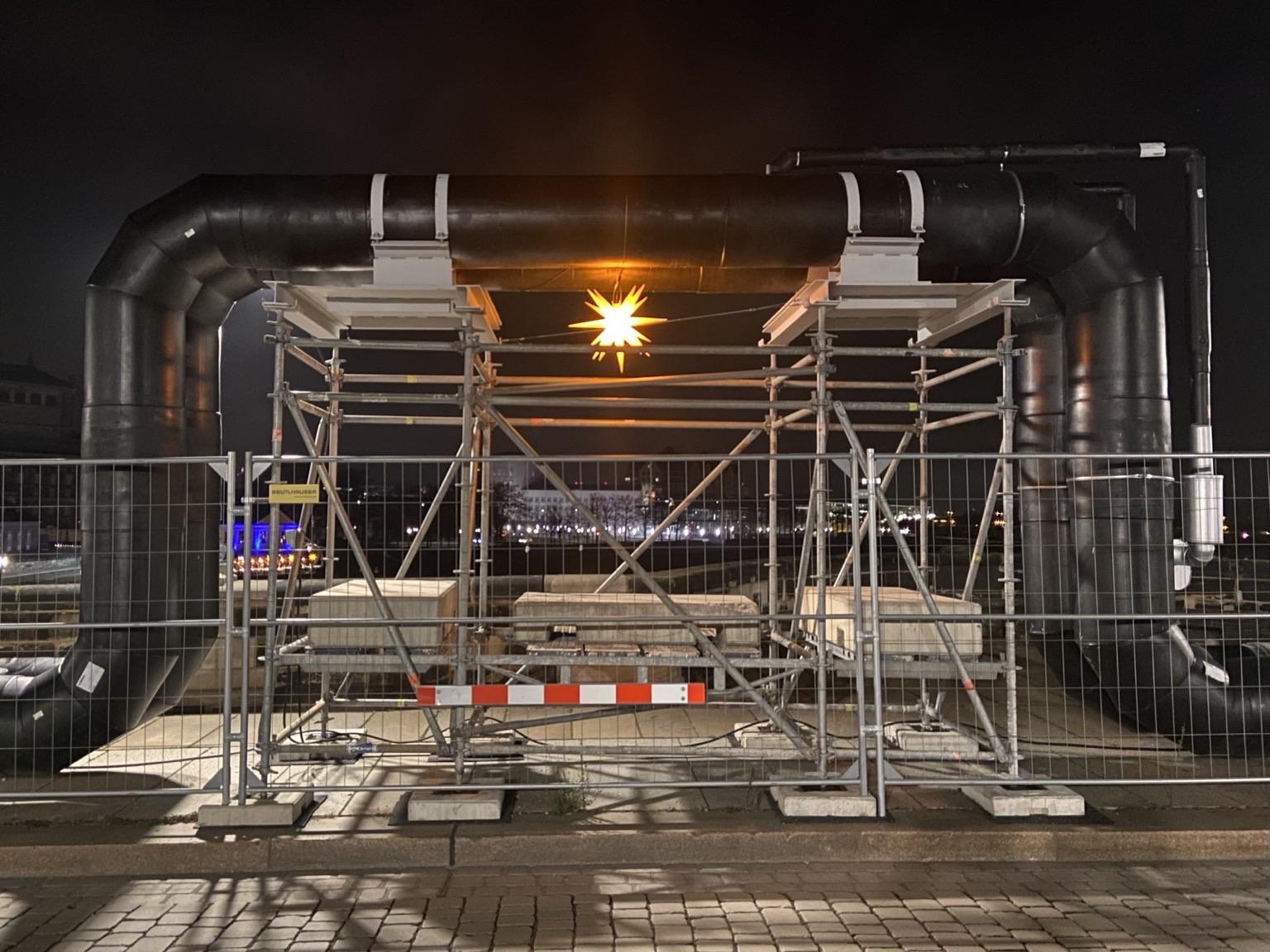 Ein Dehnungsbogen einer Fernwärmeleitung hinter einem Bauzaun. Ein leuchtender Herrnhuter Stern hängt unter dem Bogen. 