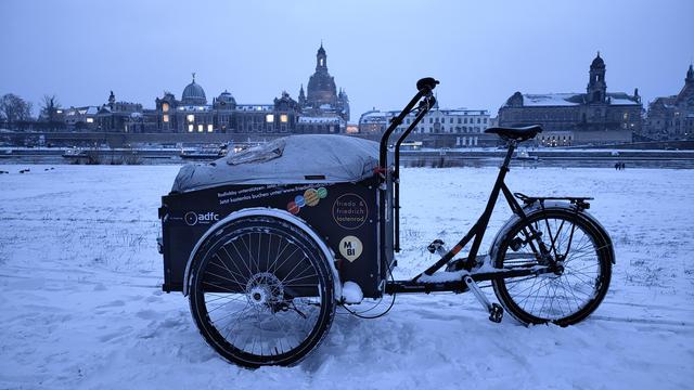 Ein dreirädriges Lastenrad vor der verschneiten Stadtsilhouette Dresdens. Der Himmel ist bedeckt und grau. 