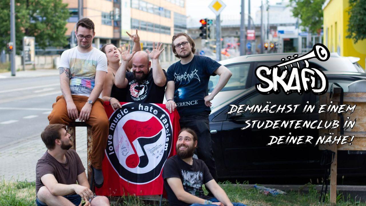 Die Mitglieder der Band SkafD sitzen um eine rote Flagge. Sie sind fröhlich und schneiden Grimassen.