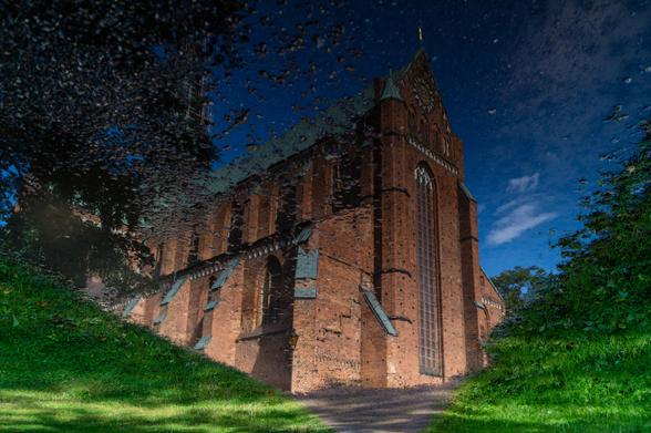 Aufnahme einer Backsteinkirche. Auf den zweiten Blick erkennt man, dass es sich um die Reflektion der Kirche in einer großen Pfütze auf einem Weg im Grünen ist, welche gedreht wurde.