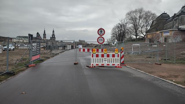 Eine Baustelle versperrt eine asphaltierte Straße mit temporären Barrieren und Verkehrsschildern unter einem bedeckten Himmel. Weiße und rote Streifenbarrieren mit orangefarbenen Warnlichtern zeigen ein Fußgängerzeichen (schwarze Fußgängerfigur in einem roten Kreis) und ein Fahrradzeichen (roter Kreis mit einem Fahrradsymbol). Ein Metallzaun umgrenzt den Bereich, wobei im Hintergrund Gebäude mit Türmen und Graffiti zu sehen sind. Eine Werbetafel auf der linken Seite zeigt die Texte "PSP" und "F…