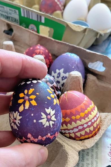 Nahaufnahme einer Hand, die ein dunkelviolett gefärbtes Osterei mit feinen floralen Mustern in Gelb, Weiß und Rosa hält, die mit sorbischer Wachstechnik erstellt wurden. Dahinter ein offener Eierkarton, in dem sich weitere bunt verzierte Eier befinden, darunter rote und lilafarbene mit dekorativen Mustern. Im Hintergrund sind weitere ungefärbte Eier im Karton zu sehen sowie eine Tischoberfläche mit Bastelmaterialien.