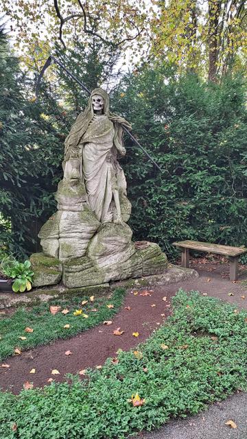 Steinskulptur des Sensenmann. Ein Skelett mit Umhang, Sanduhr in der Hand und Sense auf der Schulter. 