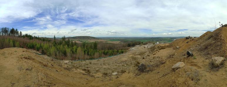 Panoramaaufnahme einer hügeligen Landschaft mit einem weiten Blick über Felder und Wälder. Im Vordergrund befinden sich trockene, sandige Hänge mit verstreuten Steinen und spärlicher Vegetation. In der Mitte erstreckt sich ein Tal mit grünen Wiesen und bewaldeten Flächen, während im Hintergrund sanfte Hügel und eine entfernte Siedlung zu sehen sind. Der Himmel ist weit und von dünnen, teils wolkigen Schleierformationen durchzogen.
